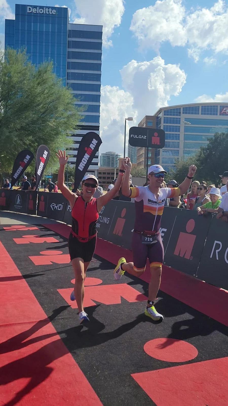 John and Annie crossing an Ironman finish line together, hands raised